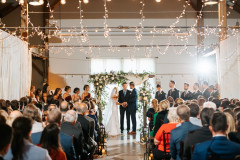 event barn hosting an indoor wedding where bride and groom are in the center surrounded by the wedding party and guests.
