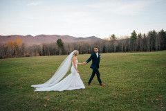 bride and groom walking in field with mountains in the background during the fall.