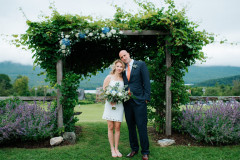 bride and groom standing in front of wedding arbor during the summer. bride holding floral bouquet.