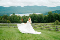 bride showing her dress and its details while standing in front of a barn fence with a lake and mountains in the background.