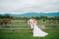 bride and groom leaning against barn fence with horses in the mid ground with the green mountains in the background.