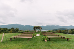 view of wedding knoll in summer showcasing floral arrangements with lanterns at the foot of multiple farmhouse benches, overlooking the green mountains.