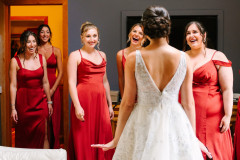 bride in white showing her dress to her five bridesmaids who are wearing red dresses. bridesmaids are all smiling and look happy.