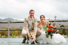 bride and groom kneeling in front of barn fence. bride is holding a red and purple bouquet. they are kneeling with their corgi, who is wearing a makeshit suit and also smiling at the camera.