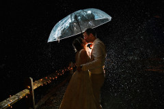 bride and groom kissing under a clear umbrella with one light behind them. kissing in the rain with a barn fence behind them.