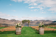Arbor setup for a wedding. Wedding Knoll features green mountains in the background and a small view of the lake below. In front of arbor are farmhouse benches on both sides creating an aisleway with two whiskey barrels at the end with floral arrangements on top of them. 