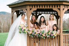 Bride standing on side of gazebo holding a floral bouquet. 5 bridesmaids are in gazebo holding flowers as well, smiling at camera.