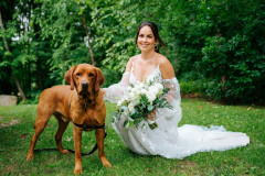 bride with bouquet with dog standing on green lawn with trees in background.