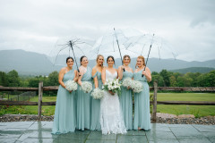 bride and bridesmaids holding umbrellas on a rainy day outside the lodge on a stone walkway.