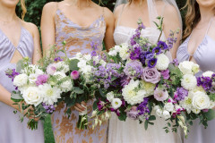 bride and bridesmaids in purple dresses holding flowers out with florals in purple, white, pink, and greenery included.