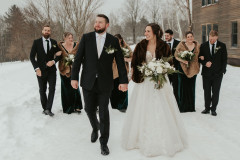 bride and groom walking towards camera with wedding party behind them. walking in the snow towards the camera with a brown building on the right hand side of the image.