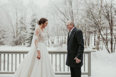 bride in white dress standing with her father for a first look with a grey fence behind them and a winterscape in the background.