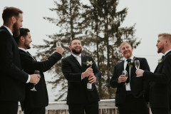 groom in middle of image with a black suit on holding a bottle of champagne, lifting to open the bottle. Groom is surrounded by four groomsmen holding glasses for champagne and smiling.