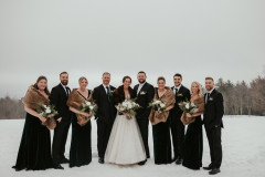 bride and groom standing in the middle with a floral bouquet with their wedding party in dark colors, also holding bouquets with white florals and greenery.