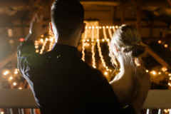 bride and groom facing away from camera looking down from loft at wedding guests with string lights behind them, 