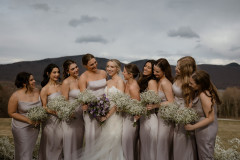 bride and bridal party standing together. bridal party is wearing light purple dresses with baby's breath bouquets and bride is wearing white, in the center, with a purple bouquet.