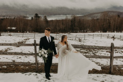 bride and groom standing in front of wooden fence with mountain scape in background and snow on the ground.