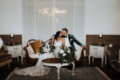 bride and groom sitting on velvet couch with floral arrangement kissing in front of sign that says "The Joneses"