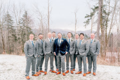 groom in blue velvet jacket with grey pants and eight groomsmen in grey suits standing on snow with boutonierres on their chests.