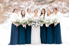 bride in white dress and six bridesmaids in blue dresses with white shawls all holding blue and white floral bouquets.