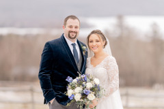 bride in white dress standing with groom in blue velvet suit holding flowers in front of a snow scape.