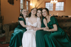 bride and bridesmaids on a couch being embraced while smiling at the camera.