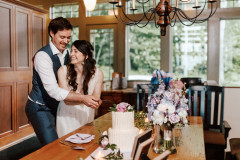 bride and groom inside home cutting a cake with flowers on table and large windows in background.
