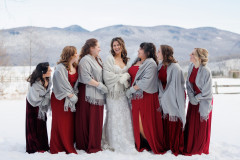 bride and bridesmaids. bride in white with white mittens and bridesmaids in red dresses with grey shawls and grey mittens surrounding bride.