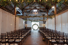 interior of event barn featuring lighting and greenery on ceiling with drapery on sides. chairs in rows down middle with circular arbor at end.