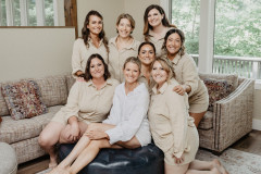 Bride in white sitting on blue ottoman. Surrounding her are seven bridesmaids in tan pajamas, all smiling at the camera. 