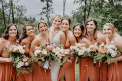 Bride in white and seven bridesmaids in a soft orange embracing each other while smiling at the camera. All of them are holding bouquets that feature pink, orange, and white florals with ferns. 