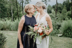 Bride in white and mother of bride in blue looking at each other. Bride is holding a floral bouquet with ferns. 