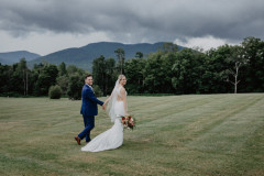 Bride in white and groom in blue suit walking away from camera but looking back at camera smiling. 