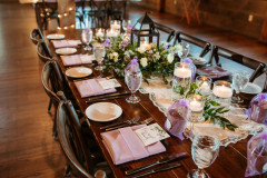 close up of table setting in event barn for a summer wedding reception featuring purple napkins, plates, and greenery in the middle of the table.