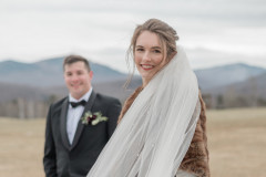 bride smiling at camera wearing a veil and a brown coat with groom in background.