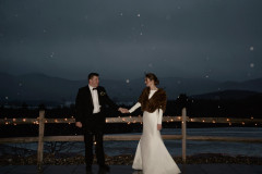 bride and groom standing in the dark in front of a wooden fence while it snows.