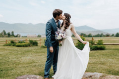 bride in white bridal gown and groom in blue tuxedo kissing on rock in front of wooden fence and mountain scape in background.