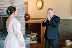 bride in white dress facing her father while he is wearing a suit, and has his hands over his mouth in surprise.
