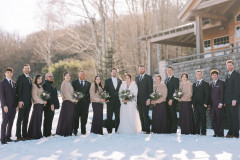 bride and groom in center of photo in white and black, surrounded by wedding party. ground is white with snow and in the background is trees along with a wooden building.