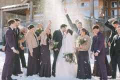 bride and groom in center of photo kissing each other while wedding party surrounds them celebrating. bridesmaids and bride are holding purple themed bouquets, and there is snow being thrown by the wedding party into the air.