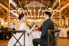 bride and groom sitting in chairs in front of camera. behind them are string lights from rafter beams and round tables with guests. bride and groom are smiling, facing each other, with glasses of champagne in hand.