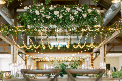 hanging ladder with white florals and greenery hanging from rafter above a sweetheart table with farmhouse chairs. background shows string lights hanging from rafters.