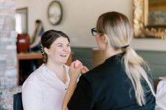 bride sitting in chair wearing white smiling while getting makeup done by a makeup artist in a black shirt with blond hair.