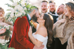 bride and groom kissing in front of camera. groom is wearing a red velvet suit jacket, bride is wearing a white dress. surrounding them are their friends smiling and celebrating.