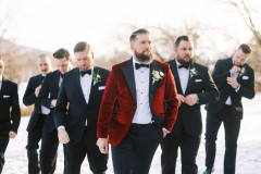 groom in red velvet suit jacket and bowtie walking towards camera. he is followed by six groomsmen in black suits and bowties. the ground is white with snow. 