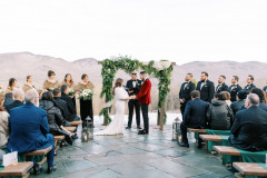 bride and groom standing at the end of the terrace patio with officiant behind them and a greenery filled arbor behind them. groomsmen and bridesmaids stand on either side while guests are seated on farmhouse benches. there is a mountain view in the background.