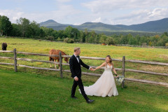 bride and groom walking in front of wooden fence with horses in pasture and mountains in background.