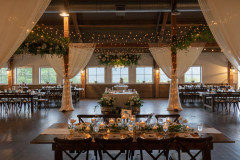 event barn set up featuring farmhouse tables, large drapes on columns and greenery hanging from lights.