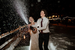 bride and groom holding bottle of champagne outside next to wooden fence and snow.
