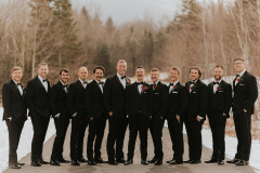 groomsmen in black suits with red boutonierres standing on road together looking at camera.